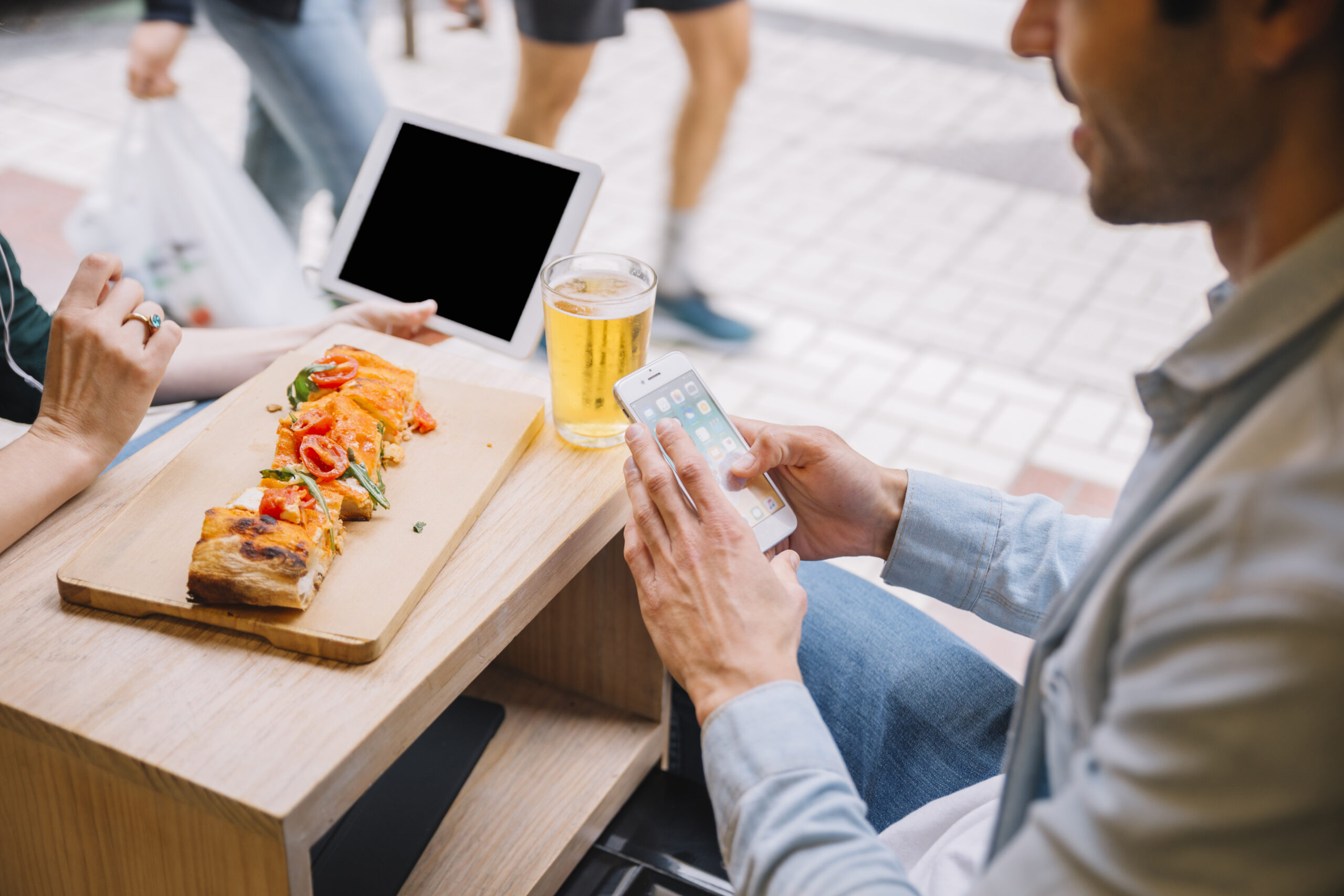 A person holding a smartphone sits at an outdoor table with a beer and a wooden board of food, while another with a tablet is partially visible in the background.